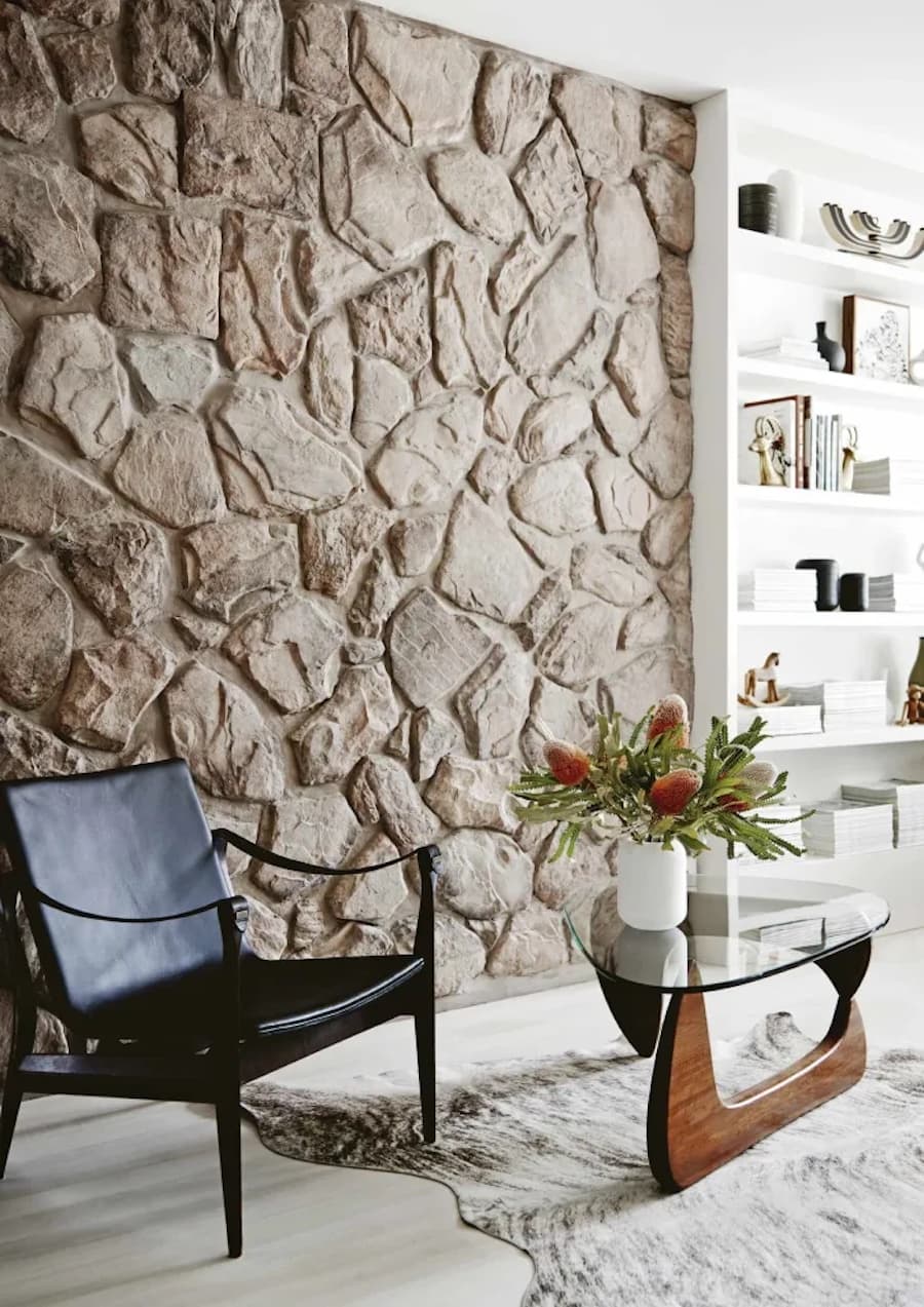 Rincón de una sala de estar con un muro de piedra junto a una biblioteca blanca con libros y adornos en sus repisas. Delante de la pared hay un sillón negro de estructura metálica y tapiz de cuero, y junto a él, una mesa lateral de vidrio.