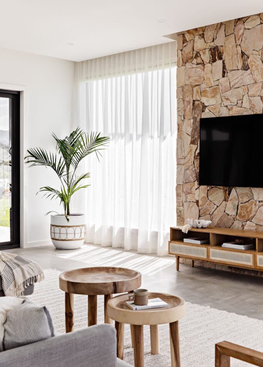 Sala de estar con un muro de piedra que sostiene un televisor. Debajo de él hay un rack con adornos y libros. Al lado del muro hay un ventanal con una gran cortina tipo visillo que deja entrar la luz natural.