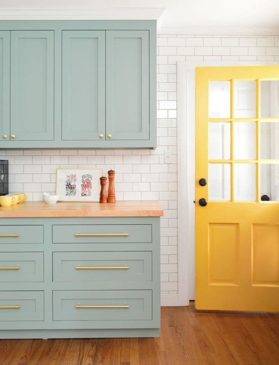 Cocina con muebles color menta y tiradores alargados de color dorado. Muebles inferiores con cajones, y los superiores con puerta. Mesón de madera clara. Muro con azulejos blancos y puerta vidriada abierta, de color amarillo.