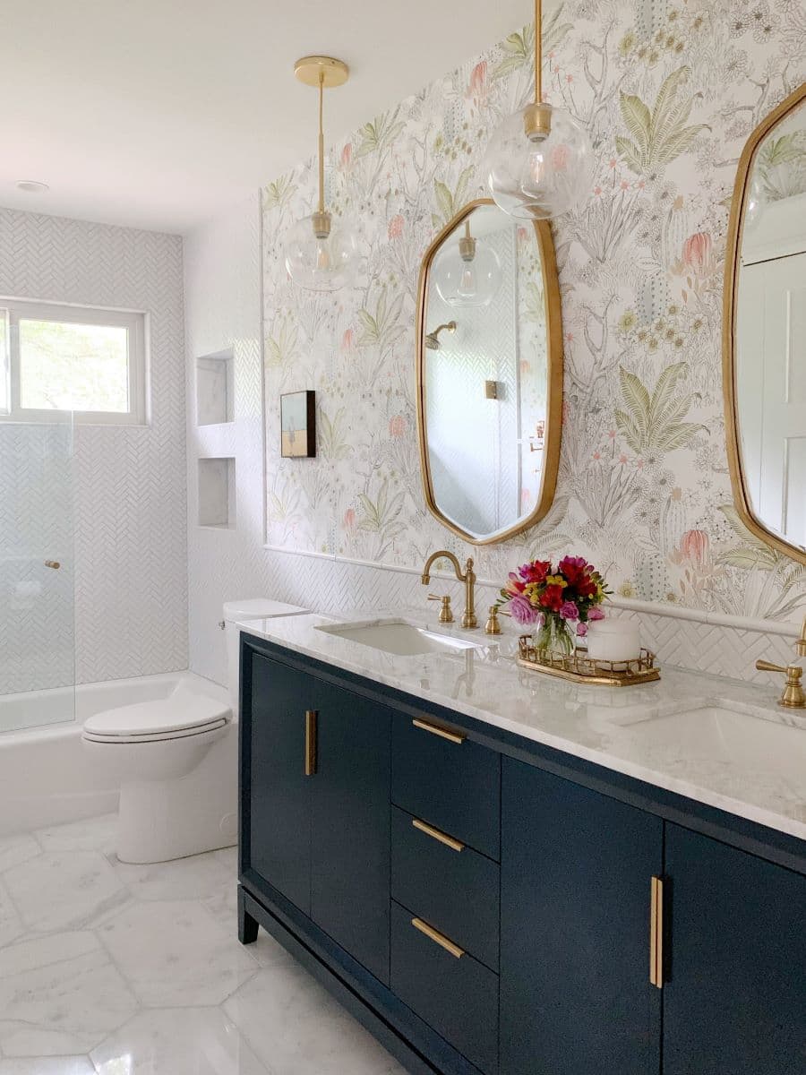 Baño de estilo clásico con papel mural de diseño floral y tropical, instalado en la parte superior de un muro. Vanitorio doble con mueble de color azul y tiradores dorados. Espejos curvos con marco dorado