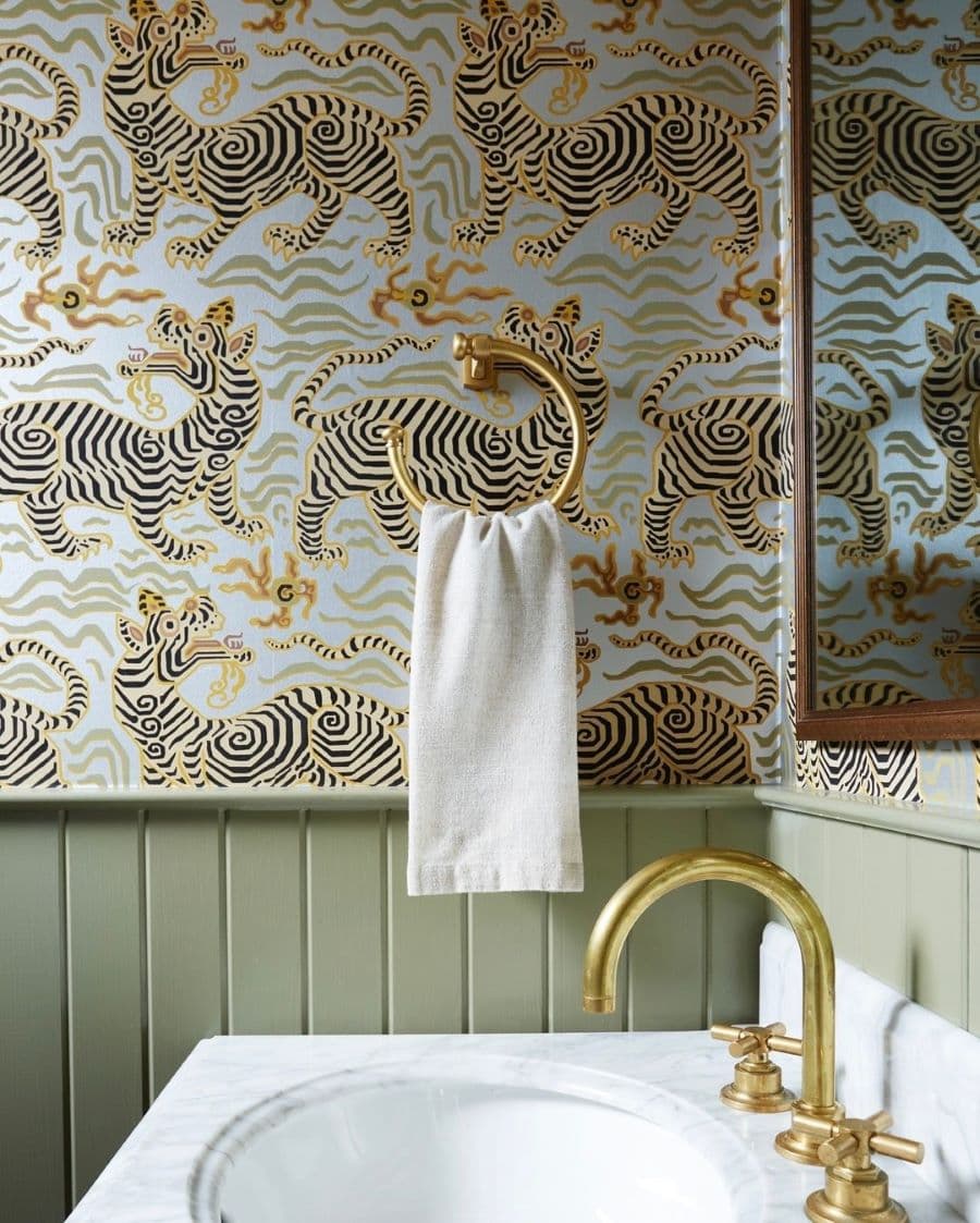 Baño con papel mural con diseño de tigre oriental. Muro con enchapado de madera verde. Porta toalla redondo, dorado, con una toalla de mano blanca.