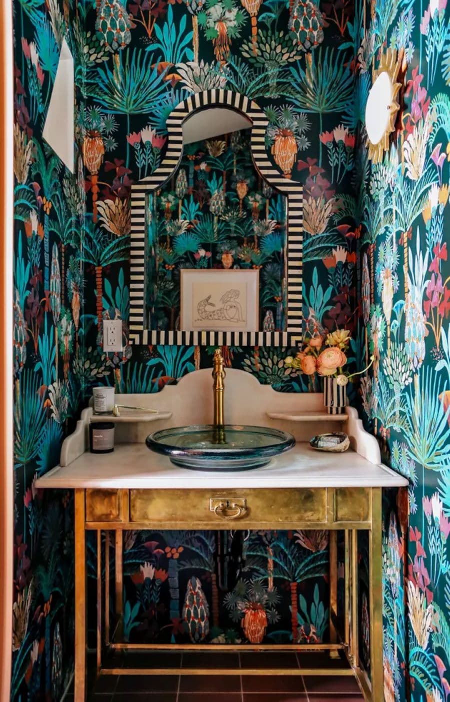 Baño de visita pequeño, con papel mural tropical oscuro instalado en todas las paredes. Vanitorio tipo mueble vintage con lavamanos de vidrio y grifería dorada. Espejo con marco con líneas blancas y negras.