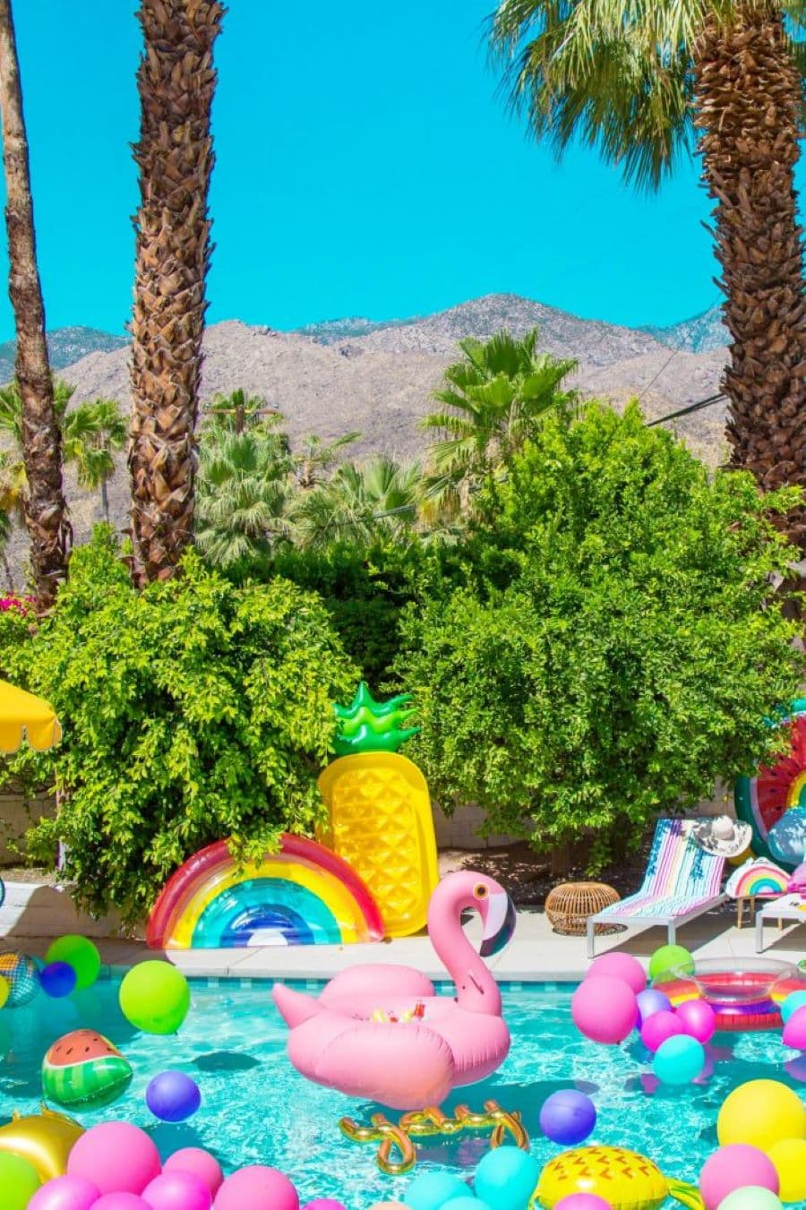 Gran piscina con globos y un flotador con forma de flamenco. Fuera de agua hay otro flotador con forma de piña y otro de arcoiris. Hay reposeras de colores y un quitasol amarillo. Atrás hay muchos árboles y palmeras.