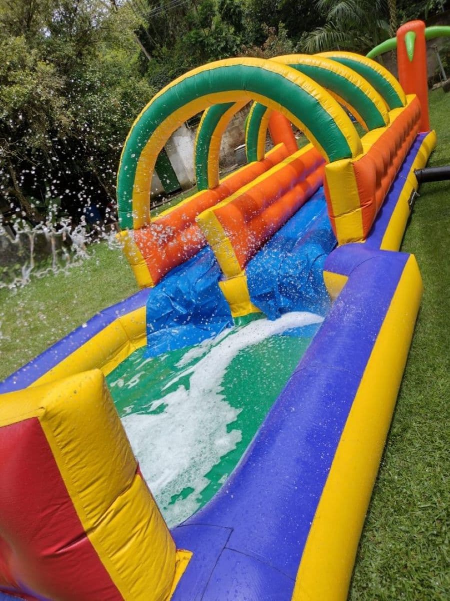 Juego de agua inflable, de varios colores. Tiene un sector con arcos y otro con una piscina y aspersores. Está sobre el pasto en un gran jardín.