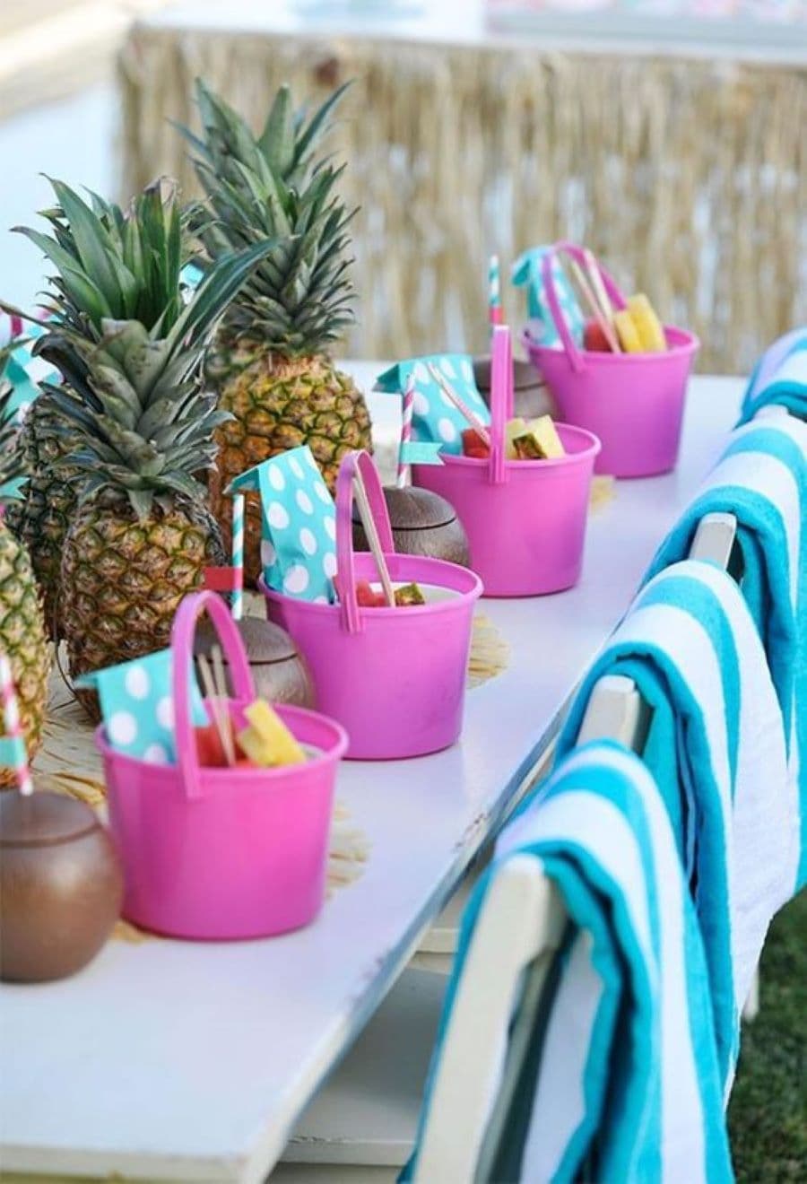 Detalle de una mesa para niños. Es blanca y arriba tiene baldes rosados con dulces. Al centro hay piñas. Sillas blancas con toallas de playa celeste y blanco.