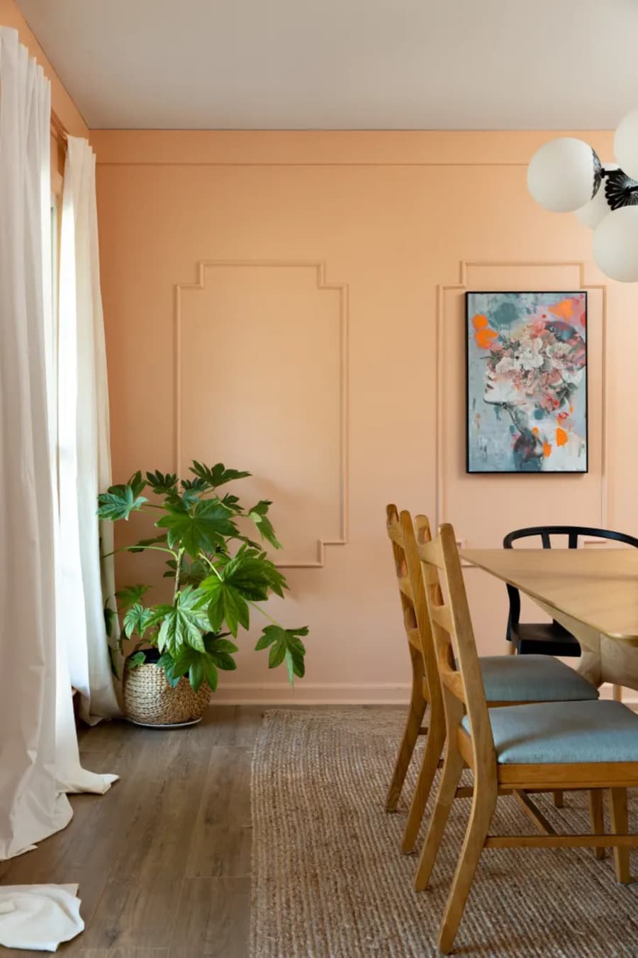 Comedor de estilo clásico, muros pintados de color Peach fuzz con molduras creando marcos. Mesa de comedor de madera con dos sillas de madera y un sitial negro. Planta en una esquina en macetero de mimbre. Ventanal con cortinas blancas