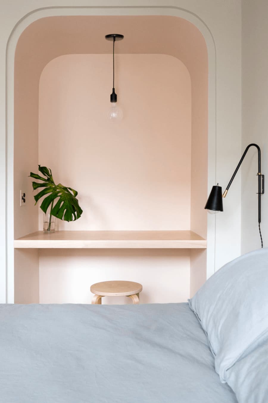 Dormitorio con closet transformado en escritorio, con una mesa flotante y un piso de madera. El espacio del escritorio está pintado de color Peach fuzz y el resto de la habitación es blanca. Sobre el escritorio cuelga una lámpara con ampolleta a la vista. 