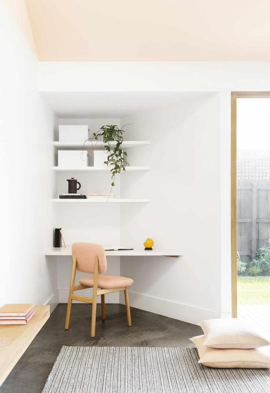 Home office de estilo moderno. Mesa flotante en una esquina con tres repisas flotantes y una sella de madera y tapiz Peach fuzz. Muros blancos techo de color Peach fuzz. Al costado hay un gran ventanal sin cortinas. Piso de concreto gris oscuro