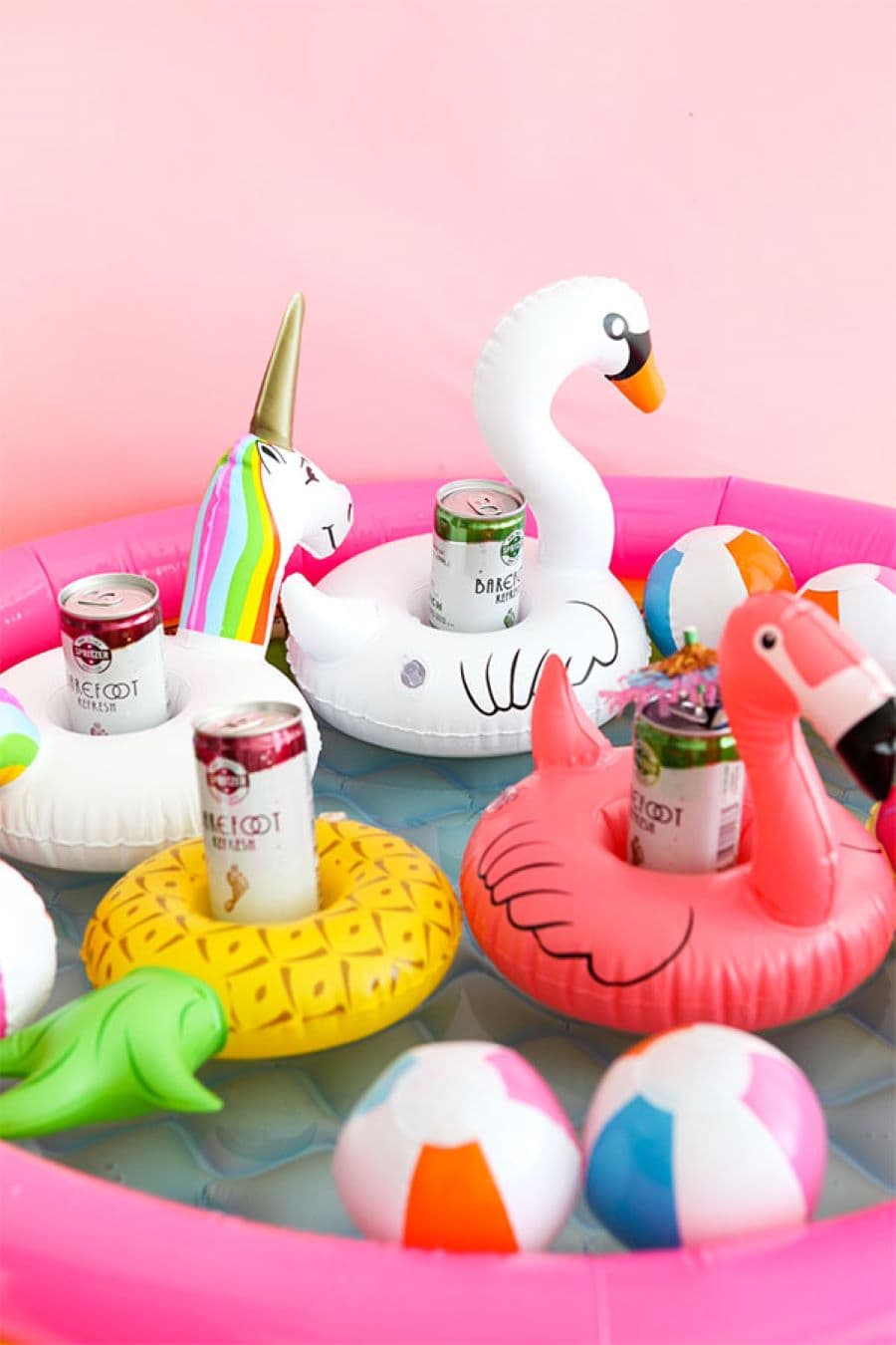 Detalle de una pequeña piscina inflable, con mini flotadores posavasos, con formas de flamenco, cisne, unicornio y piña. Los posavasos sostienen latas de cerveza. Tres pequeñas pelotas inflables en color blanco, rosado, celeste y naranjo.