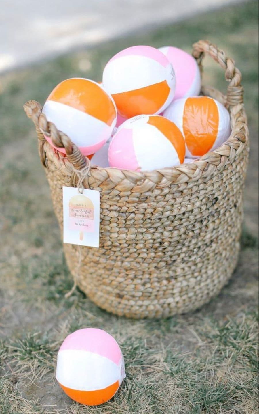 Detalle de canasto de mimbre con pelotas pequeñas inflables, de color blanco, naranjo y rosado. En una de las asas del canasto cuelga una etiqueta de papel.