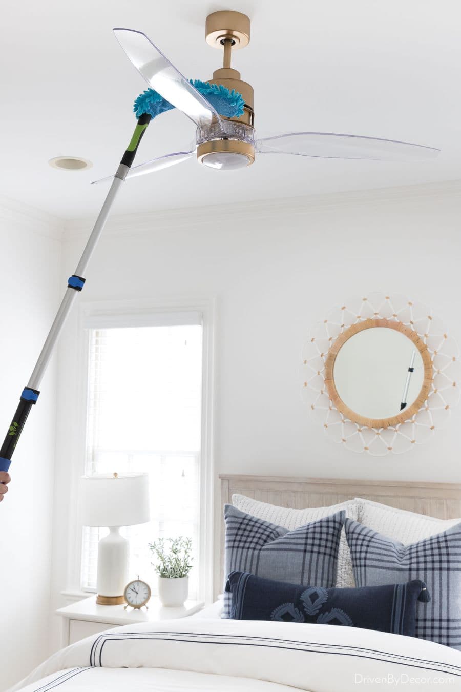 Persona limpiando un ventilador de techo con un plumero de mango extensible. Dormitorio de estilo clásico con cama de dos plazas, con textiles en colores blancos y azul. Sobre la cama hay un espejo redondo con marco dorado. Velador blanco con lámpara.