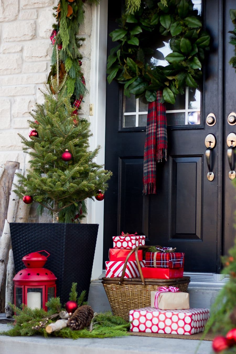 Entrada de una casa. Puerta negra con vidrio con una gran corona de navidad con una cinta escocesa. En el suelo hay varios regalos envueltos con papel y cintas. Al costado hay un pino en un macetero negro, cuadrado. Baje él hay un fanal rojo