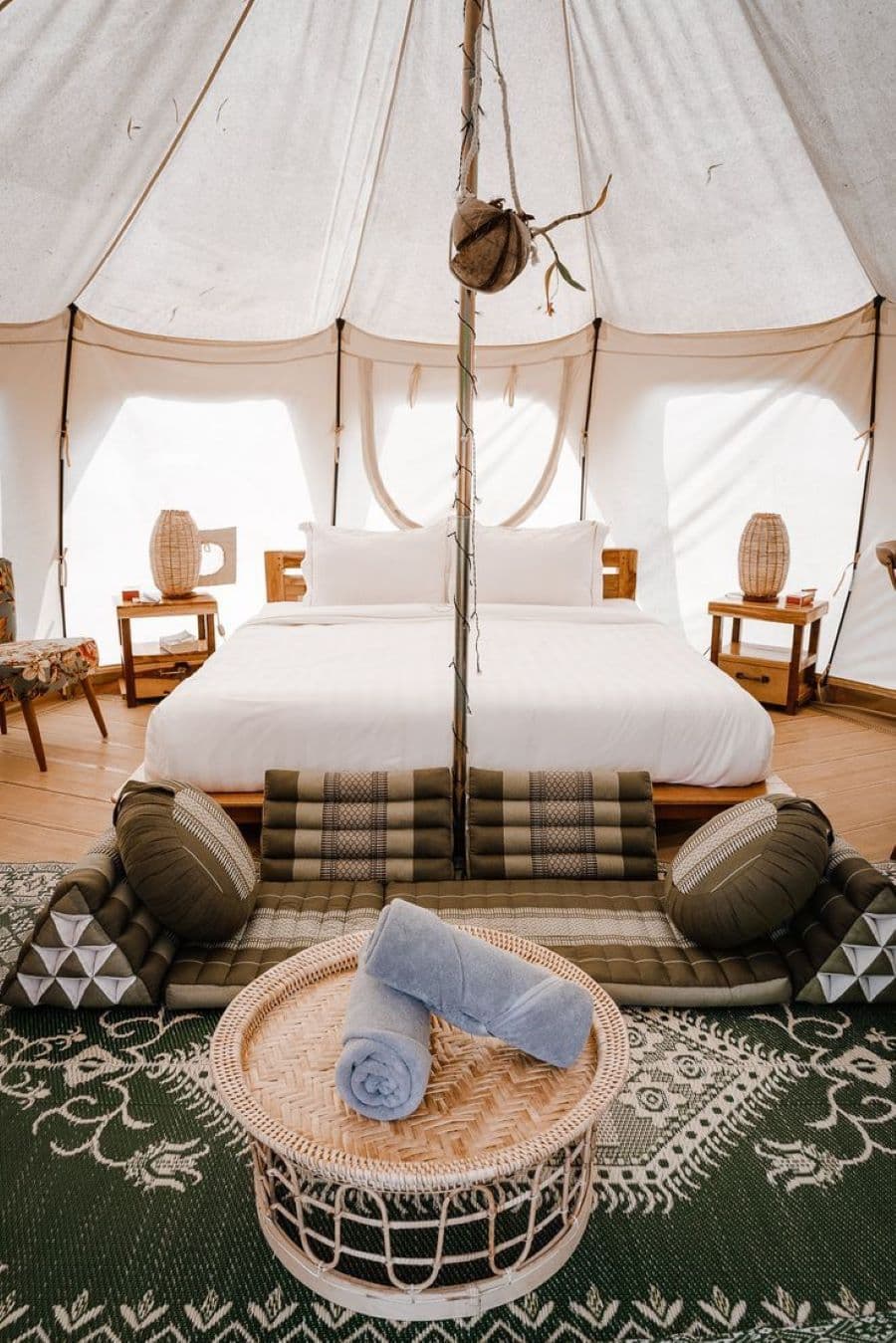 carpa de glamping blanca, muy amplia. Gran cama con base de madera y cubrecama blanco. Sillón hecho con cojines en el piso, sobre alfombra negra con diseño blancos. Mesa de centro de mimbre. Dos veladores de madera con lámparas de mimbre