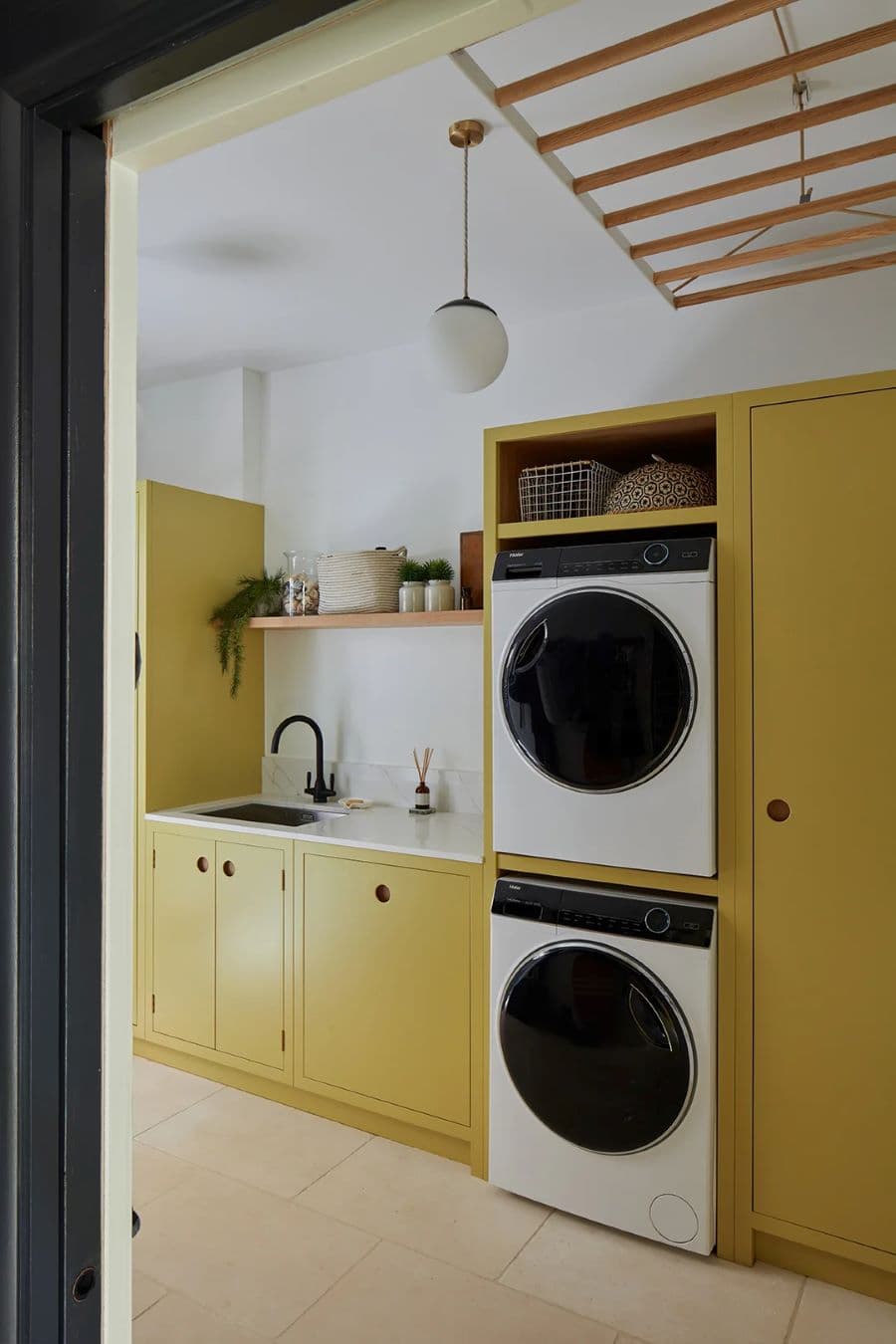 Logia con muebles a medida de color mostaza. Lavadora y secadora empotradas, una sobre la otra. Lavadero con llave negra y mesón blanco. Repisa flotante con plantas y caja organizadora. Colgador de techo de madera