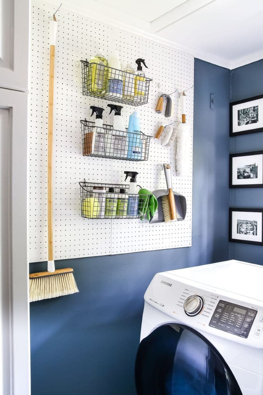 Logia con pared azul y organizador de muro blanco hecho con cartón blanco con agujeros. De él hay colgado un escobillón y canastos metálicos negros con útiles de aseo. Hay una lavadora blanca y en la pared hay colgados tres cuadros blanco y negro