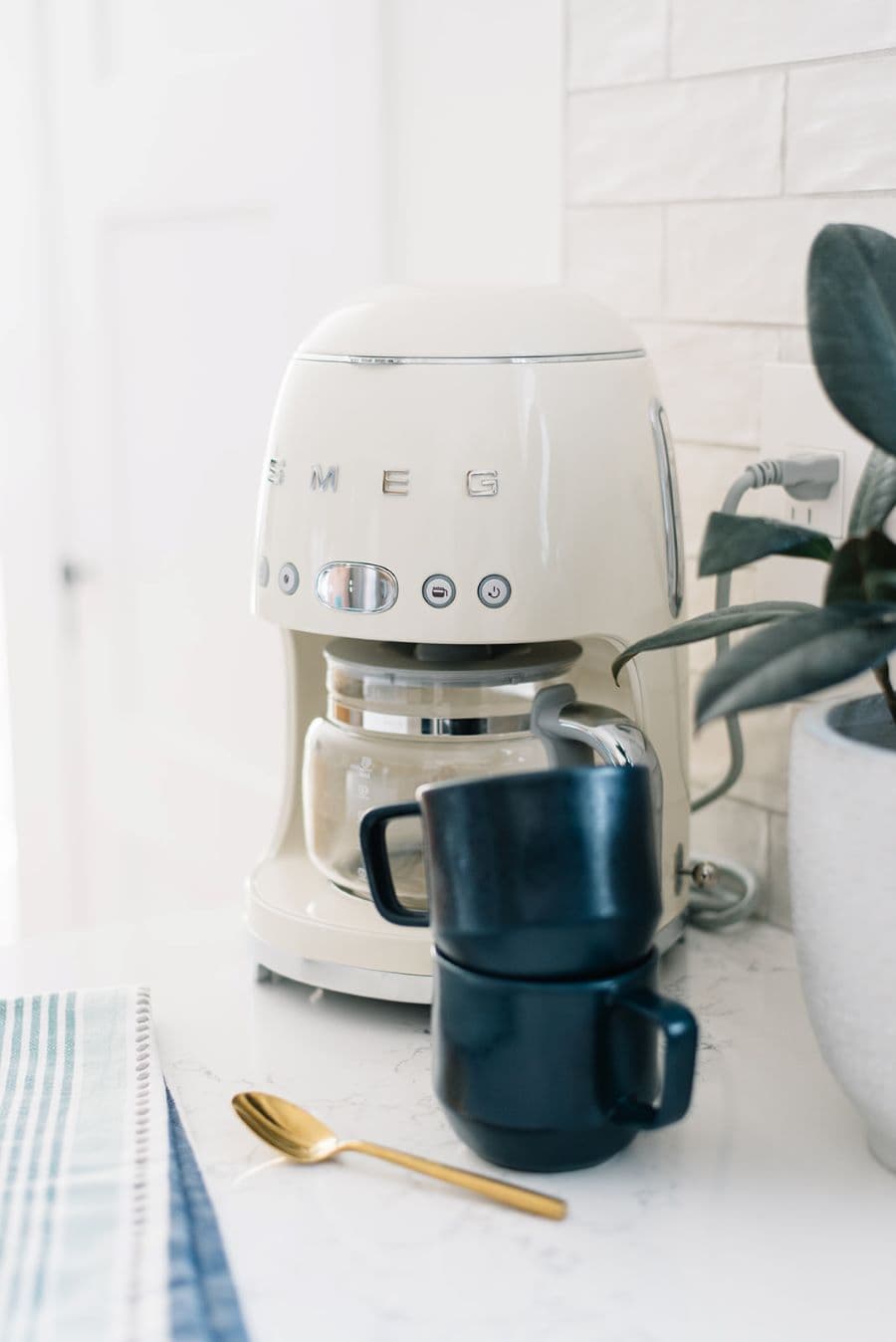 Cafetera blanca de estilo vintage, con dos tazas negras, una sobre otra, delante de ella. Pequeño gomero al costado en un macetero blanco y un paño de cocina blanco con líneas azules. Cuchara dorada. 