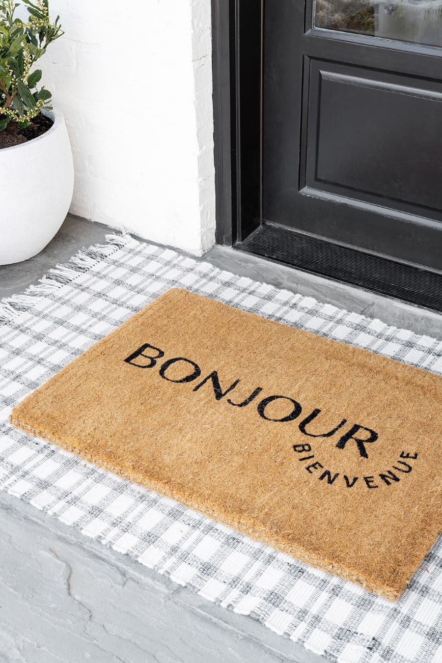 Entrada de una casa con un limpia pies que dice "bonjour" y "bienvenue" Es de color café y bajo él hay una pequeña alfombra a cuadrillé blanco con gris. La puerta es negra, el piso gris y se ve la esquina de un macetero blanco