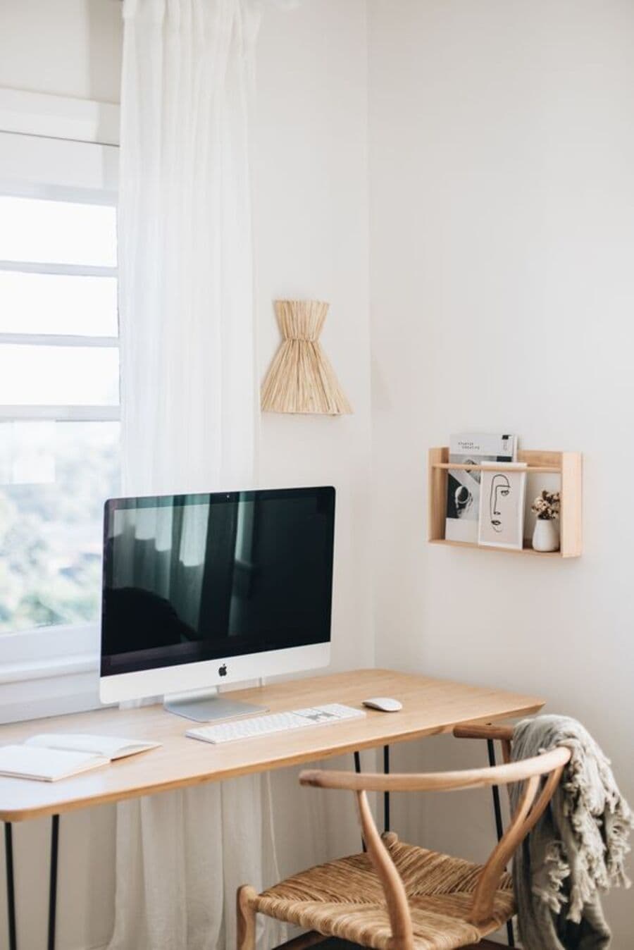 Escritorio clásico, mesa de madera, con computador imac sobre ella. Silla de madera con asiento de mimbre. Muros blancos con lámpara de muro de mimbre. Pequeña repisa de madera con revistas. Gran ventana con cortina blanca