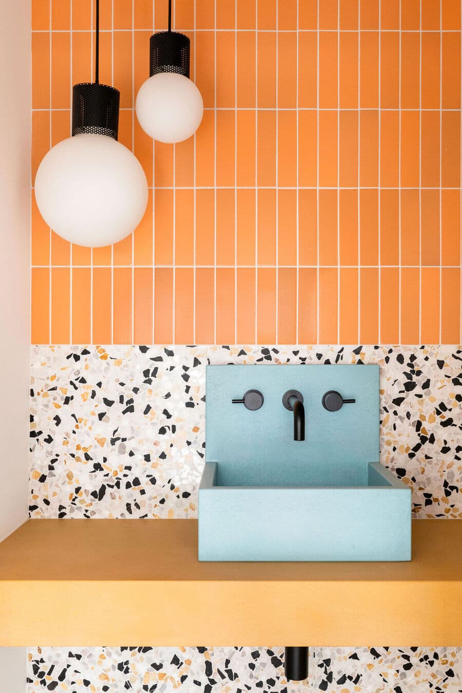 Detalle de un baño con mucho color. Azulejos amarillo anaranjado, de forma alargada. Muro de terrazo y lavamanos celeste con grifería negra. Dos lámparas de techo redondas con cable negro.