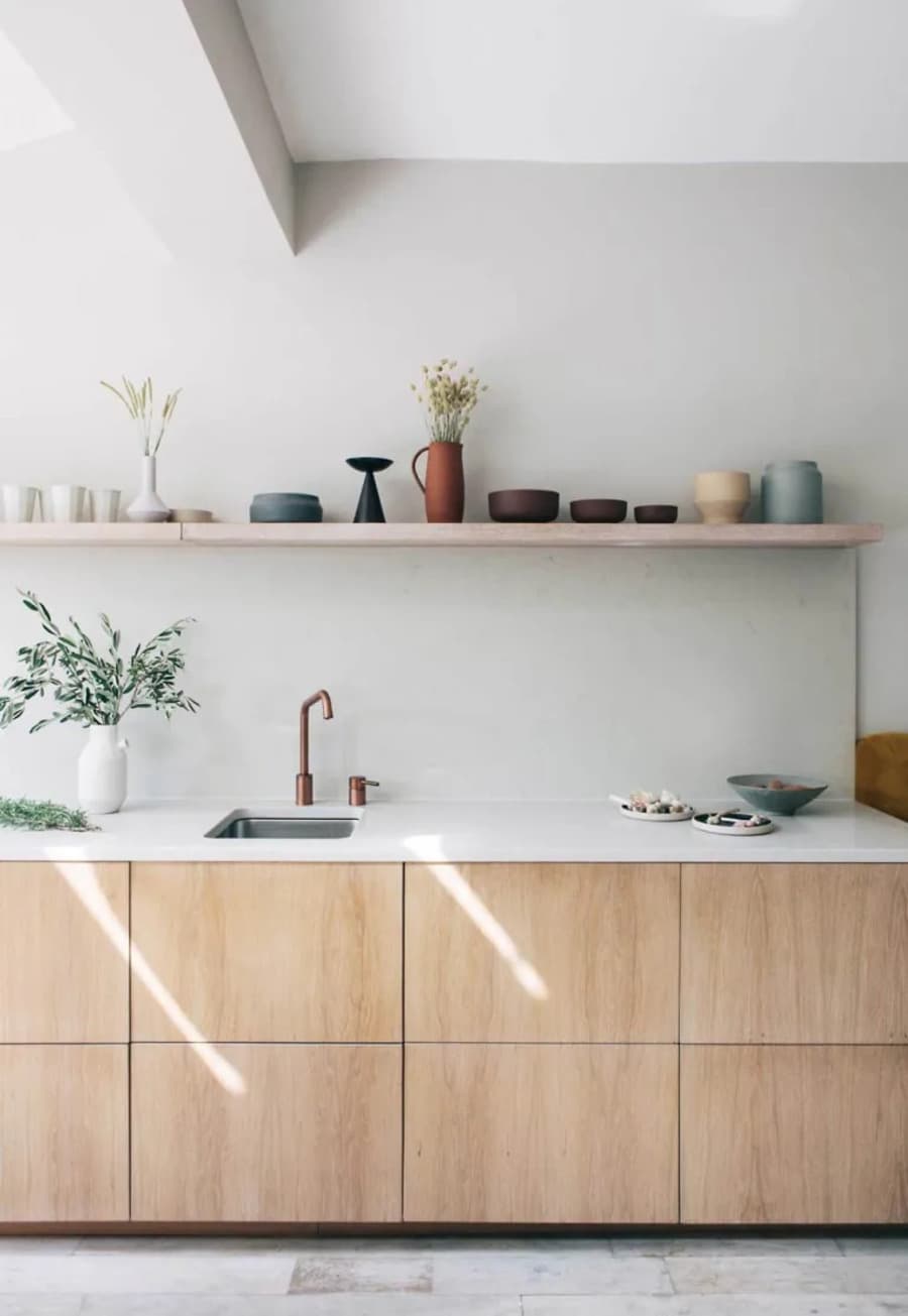 Cocina minimalista al muro. Mueble de madrea inferior, con puertas. Mesón blanco. Repisa flotante alargada con vajilla de cerámica. Muros blancos y piso de cerámica tipo piedra, gris claro.