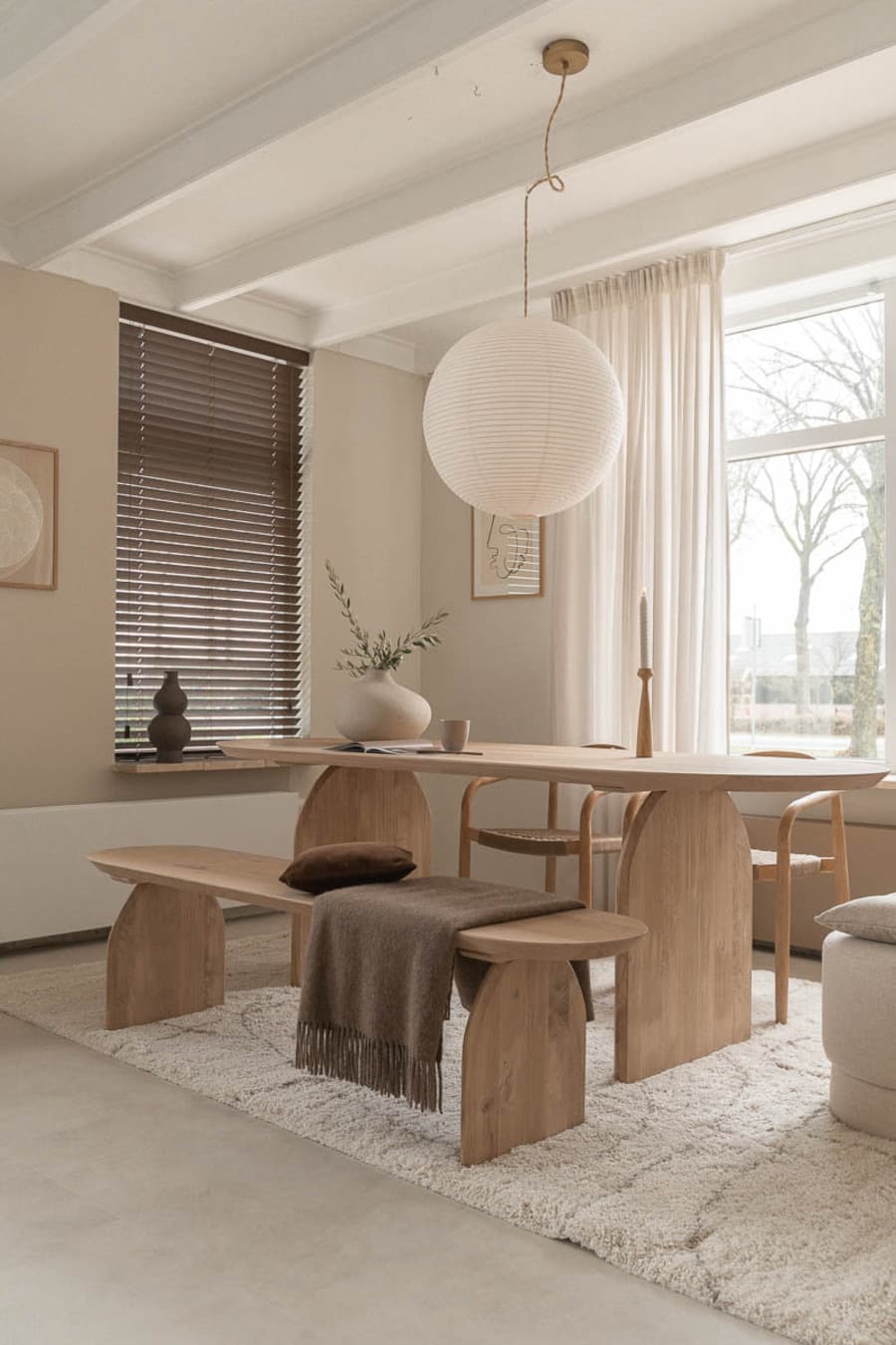 Comedor estilo japandi, orgánico. Mesa de madera, de líneas redondeadas. A una lado hay una manqueta de madera y al otro dos sillas de madera. Gran lámpara de techo de papel, redonda. Alfombra blanca, ventanal con cortinas blancas. Muros de color beige