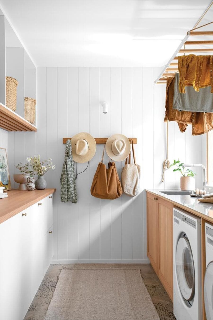 Sala de lavado de estilo boho. Mueble lateral pegado al muro con mesón de madera y puertas, con una repisa flotante blanca. Al frente un mueble a medida con la lavadora y secadora. Secador de ropa de madera, colgado del techo. Muros blancos.