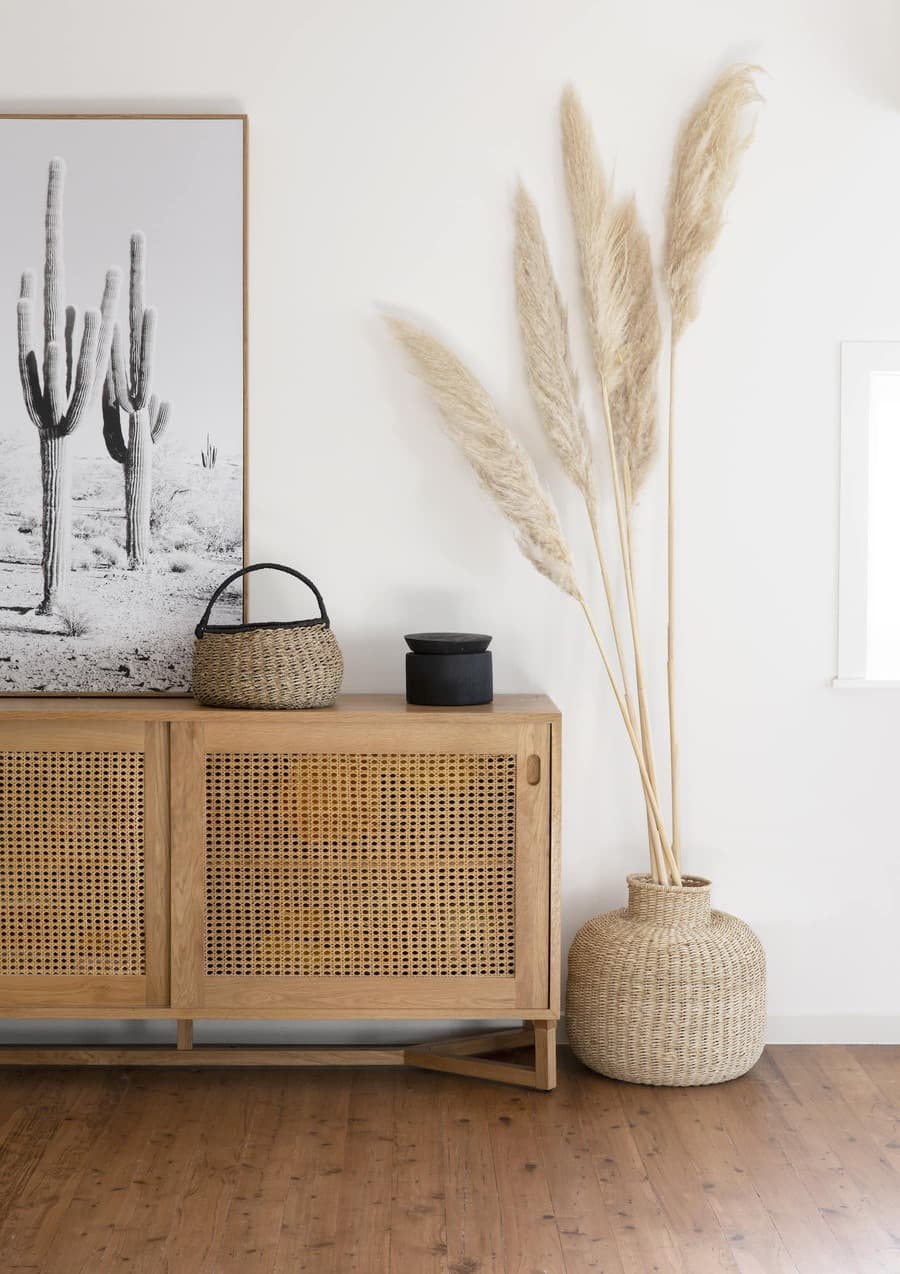 Detalle de espacio bohemio, con mueble tipo arrimo de madera con dos puertas enjuncadas. Sobre él hay un cuadro de cactus color blanco y negro, un canasto de mimbre. Al costado un macetero de mimbre en el suelo con 4 colas de zorro altas. Muros blancos.