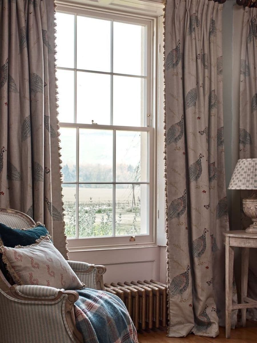 Living estilo clásico vintage, con una gran ventana con marco de madera y cortinas con estampado de faisán en tonos crema y azul, con borlas en los extremos. Sitial estilo francés con dos cojines y una manta escocesa. Al frente hay una mesa de apoyo blanca