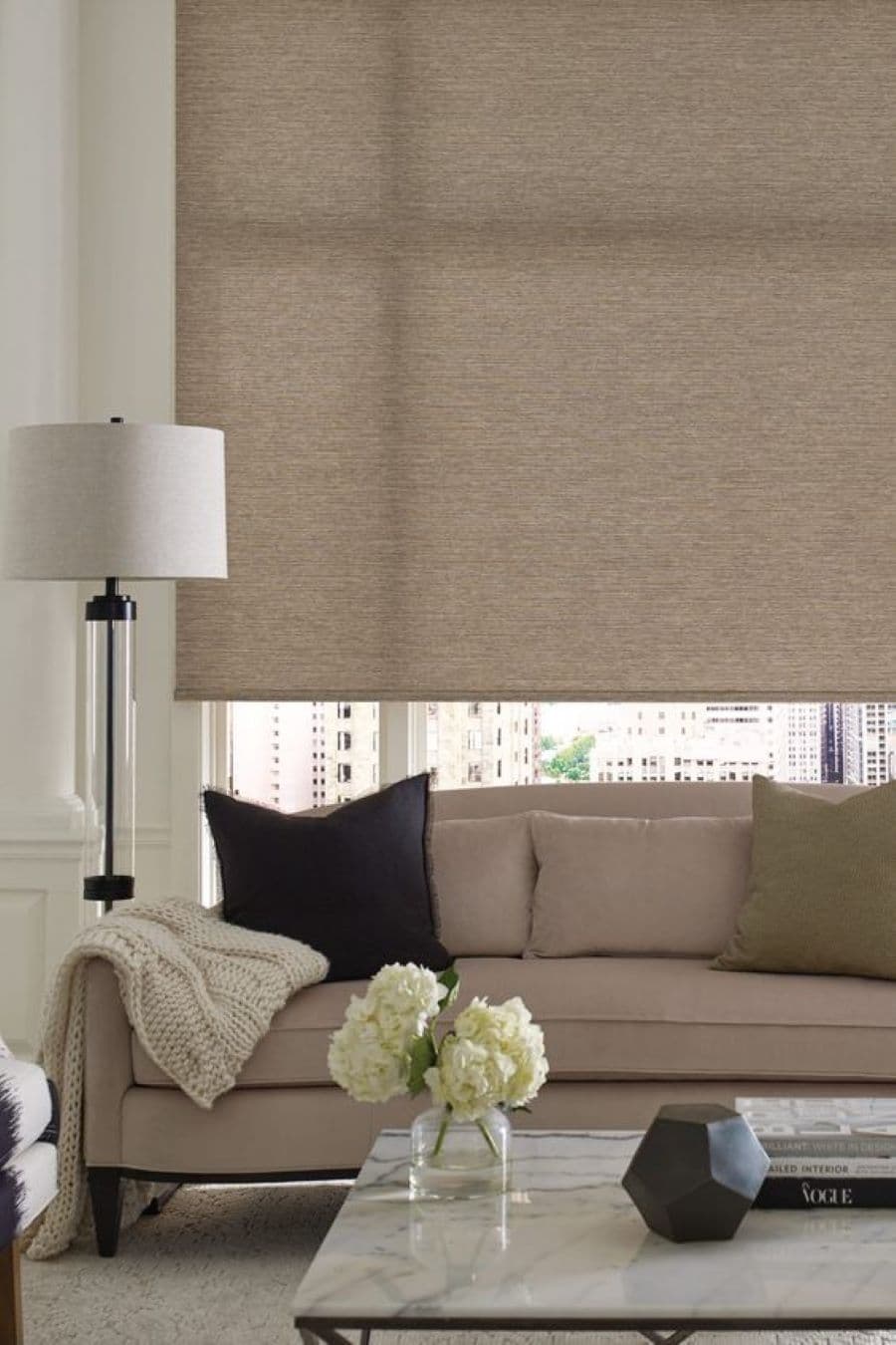 Living clásico con gran ventana con una cortina roller abajo, de color beige oscuro. Sillón clásico, beige, con un cojín negro, dos beige y uno verde, además de una manta blanca. Al costado hay una lámpara de pie de base negra y pantalla blanca redonda