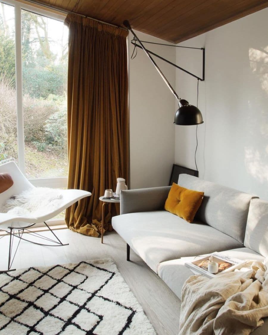 Living de estilo moderno con gran ventanal y muros blancos. Cortina de color mostaza de terciopelo, que cae sobre el suelo. Sillón gris de líneas simples, con cojín pequeño café. Sitial moderno blanco con patas negras de fierro. Lámpara de muro industrial