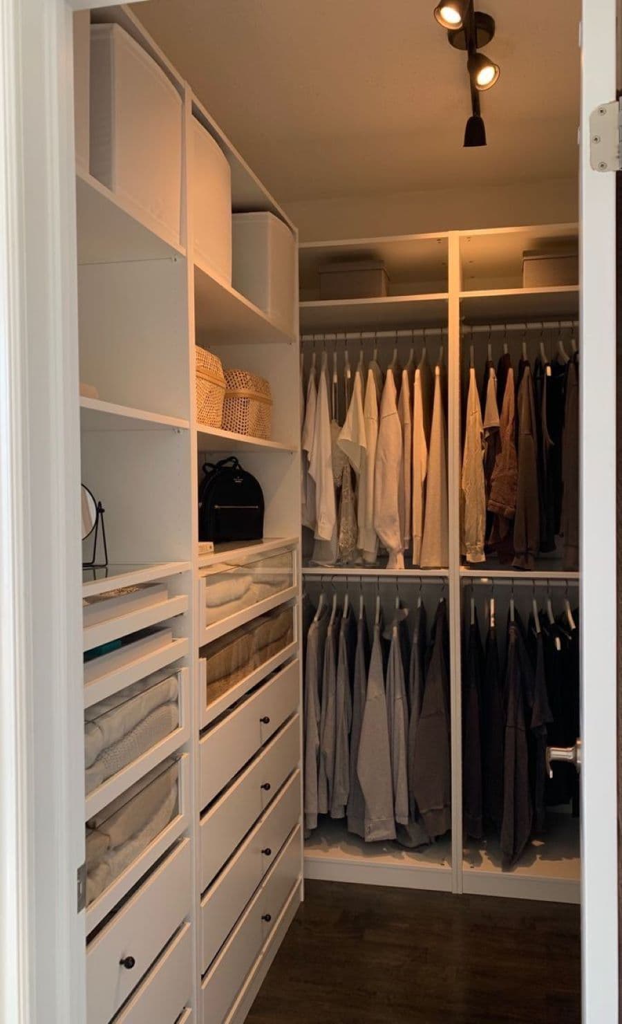 Walk in closet pequeño, con muebles de melamina en color blanco. Tiene repisas y cajones, además de colgadores con camisas y pantalones. Hay una luz de techo que está encendida y es de tonalidad cálida.