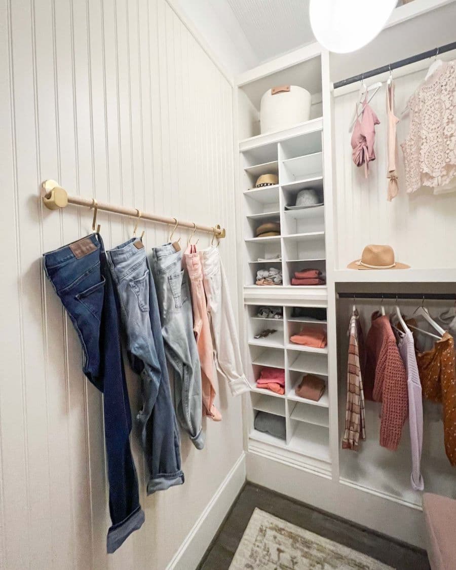 Walk in closet pequeño, de color blanco, con repisas pequeñas y dos barras con ropa colgada. En el muro del lado hay una barra con ganchos dorados donde cuelgan pantalones de diferentes colores.