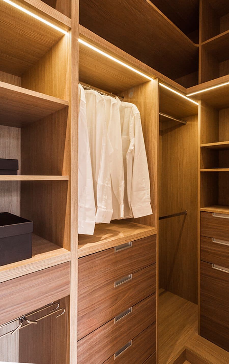 Walk in closet pequeño, con muebles de madera. Tiene repisas y cajones, y una barra con camisas blancas colgadas, Tiene luces led empotradas en la parte superior del closet, con tonalidad cálida.