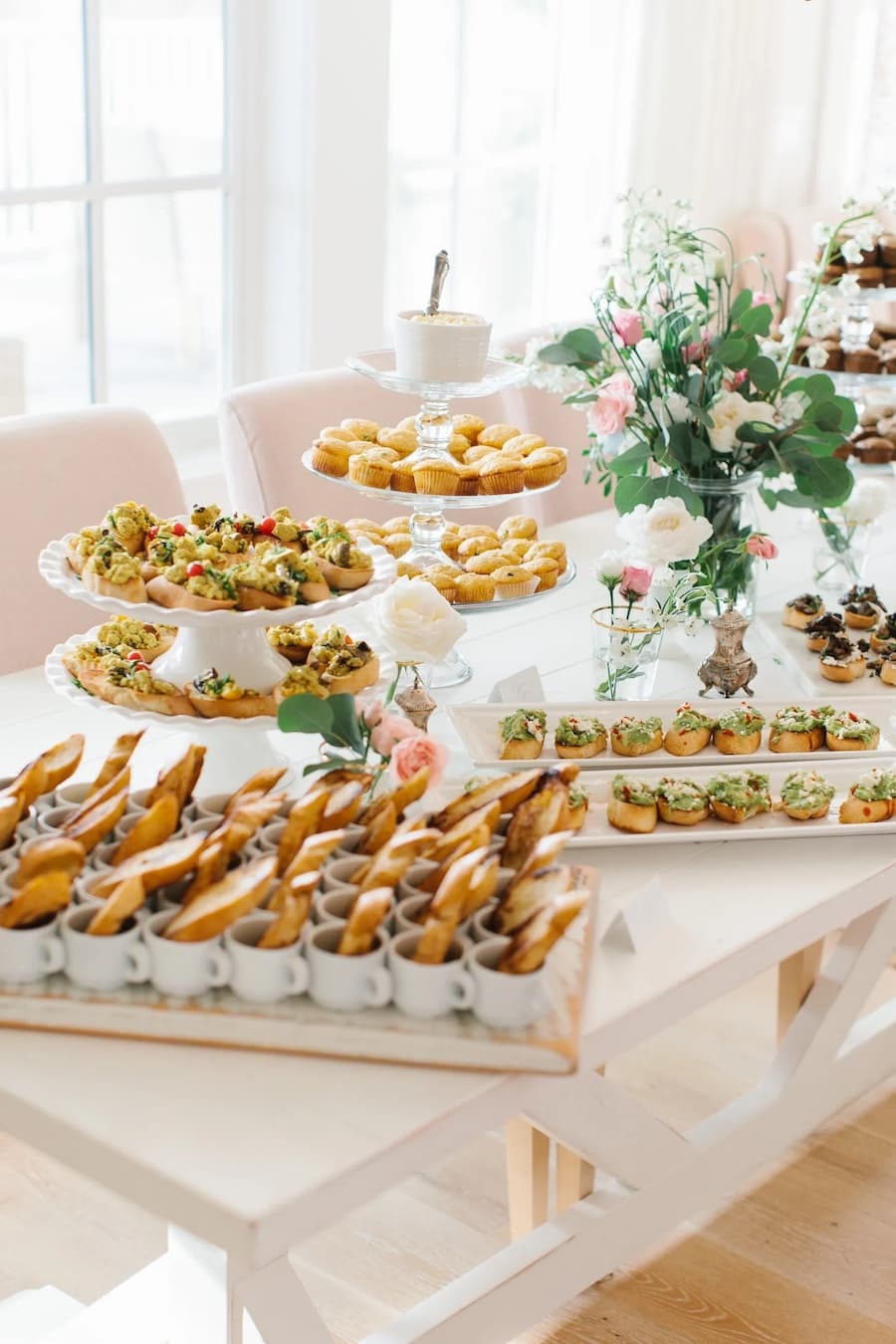 Mesa con sándwiches y snacks salados dispuestos en diferentes bandejas de color blanco. Al centro hay un arreglo floral.