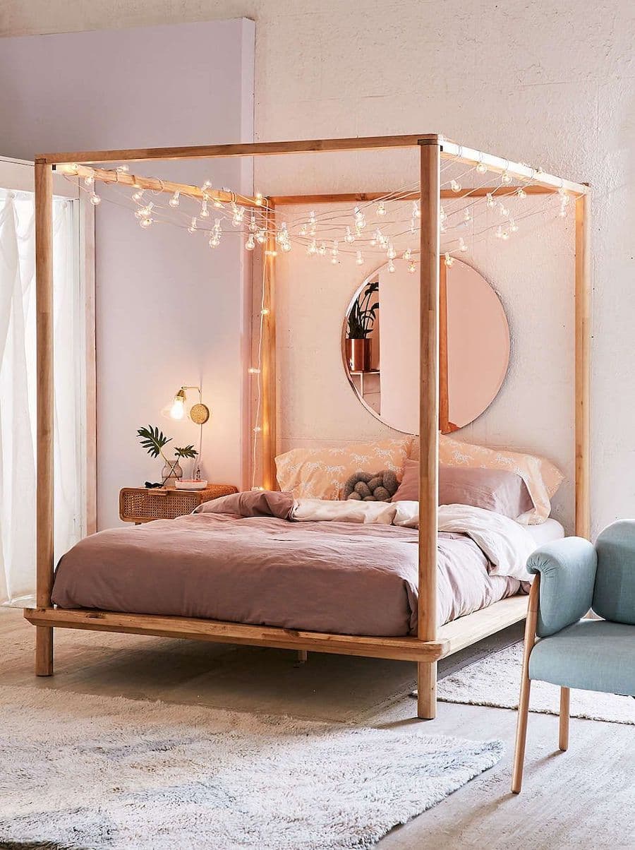 Cama con estructura de madera que sostiene guirnaldas decorativas. Como respaldo de cama hay un gran espejo redondo. 
