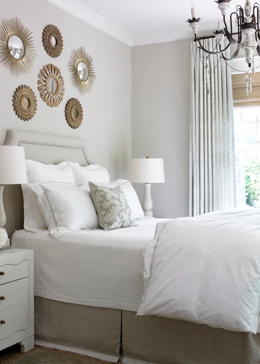 Dormitorio de tonos blancos con una serie de mini espejos con marco dorado y diseño estilo sol sobre el respaldo de la cama.