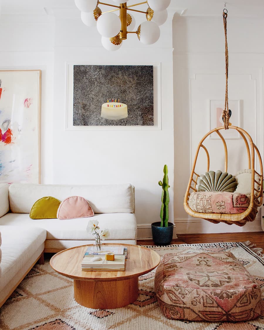 Sala de estar de estilo boho chic con una alfombra con diseño en tonos tierra, un sofá seccional blanco con cojines en colores pastel, una mesa de centro redonda de madera y una silla colgante de mimbre con cojines que combinan con la alfombra.