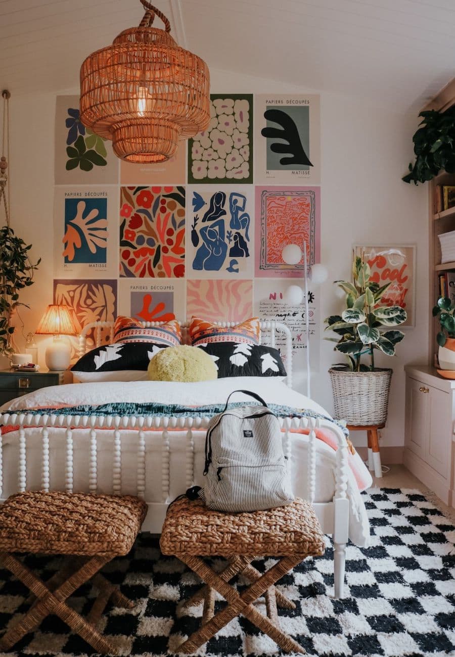 Dormitorio aesthetic con una cama de 2 plazas de estructura blanca, cojines y piecera de colores. En el muro sobre el respaldo hay una serie de ilustraciones de colores, la bajada de cama tiene un patrón de ajedrez en blanco y negro.