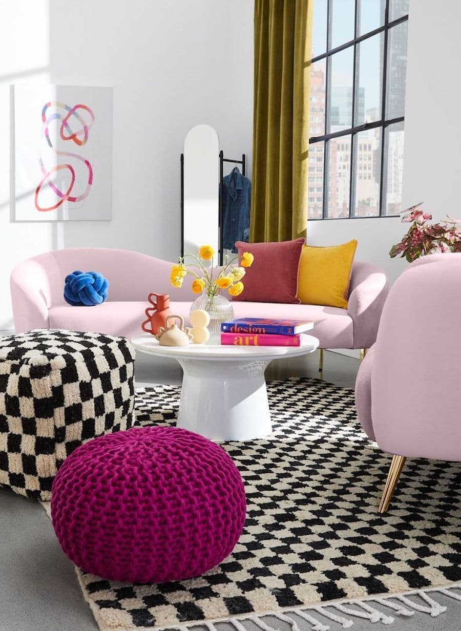 Living colorido con sofás rosado pastel con cojines de colores, un pouf fucsia y un pouf con estampado de ajedrez en blanco y negro. La alfombra tiene el mismo diseño que el pouf.