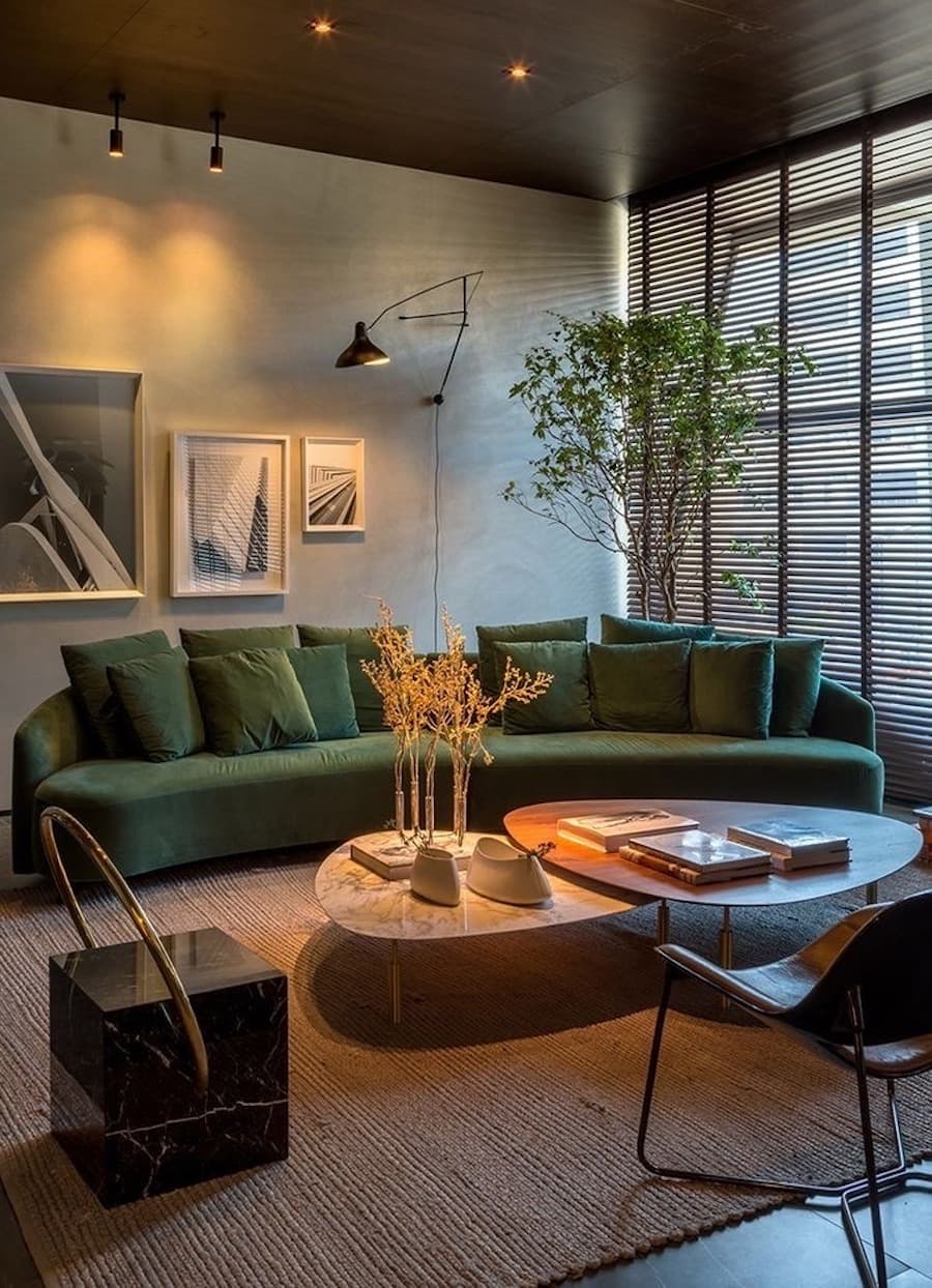 Sala de estar con un gran sofá verde oscuro y dos mesas de centro que se sobreponen. Hay distintos tipos de iluminación, como focos y una lámpara de pie, que crean una cálida atmósfera.
