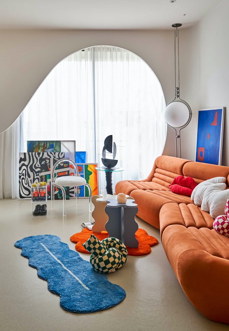 Sala de estar de estilo retrofuturista con un sofá anaranjado, decoraciones con forma de ondas, cuedros en el suelo y otros adornos modernos.