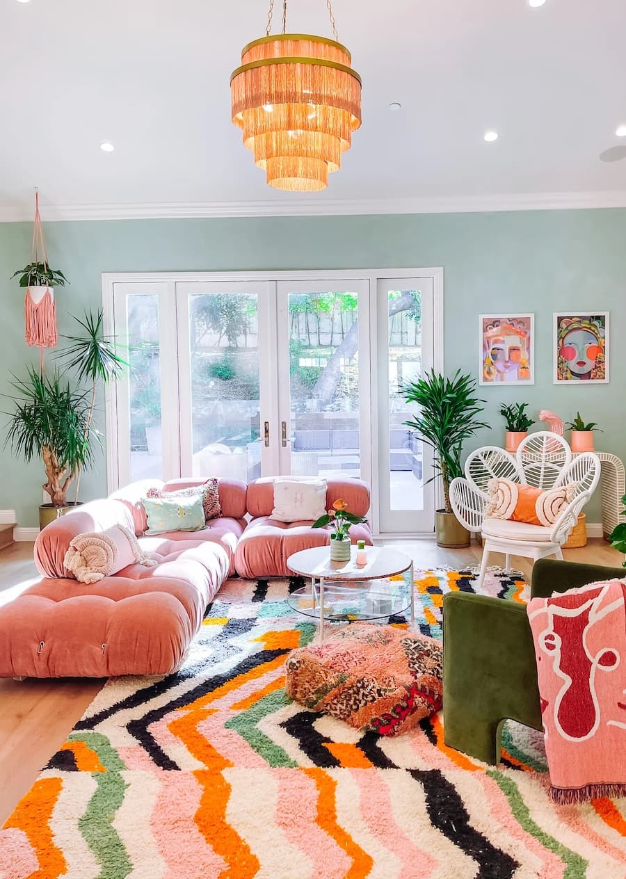 Sala de estar con estilo setentero con una gran sofá seccional rosado, una alfombra de líneas de colores, cojines y plantas en el suelo.