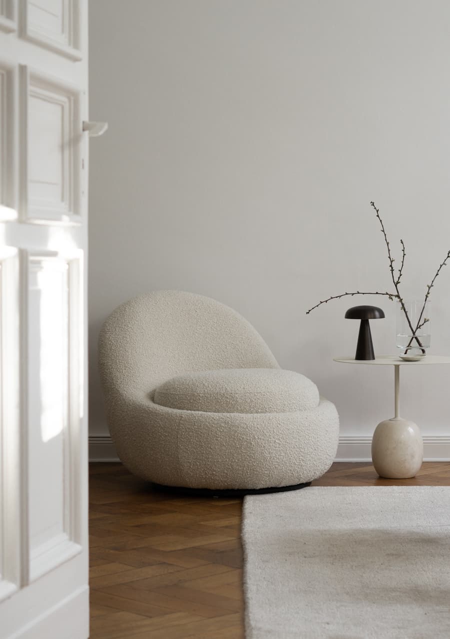 Rincón de una sala de estar con un sillón de 1 cuerpo con tejido de lana bouclé blanco, junto a una mesa lateral escultórica con cubierta de vidrio circular. También hay una alfombra blanca.