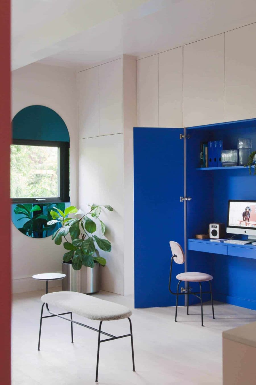 Habitación moderna, con closet en todo un muro, con puertas blancas. El closet está abierto y dentro es azul cobalto. Tiene un escritorio escondido, con una mesa empotrada y una repisa flotante. Silla negra de fierro con tapiz rosa. 