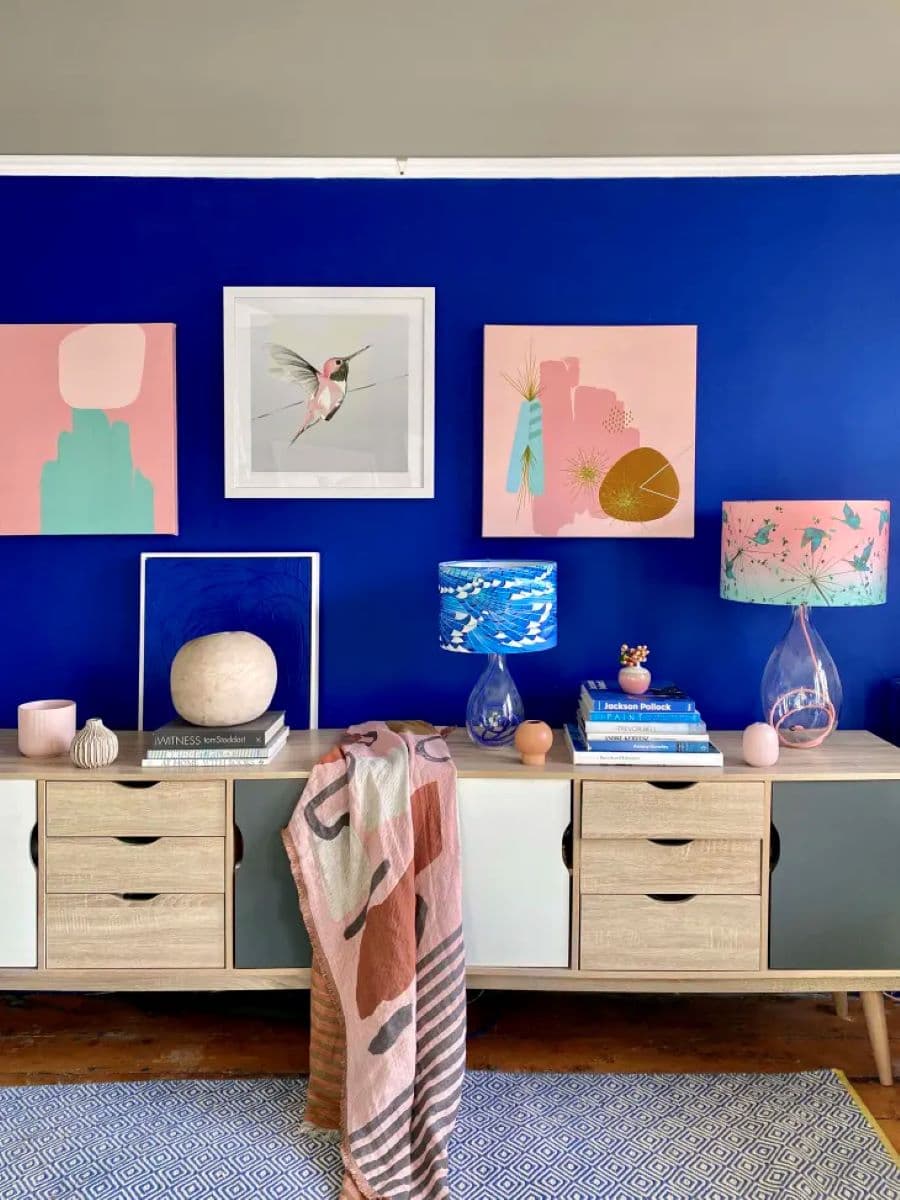 Living con pared pintada de azul cobalto. Muro galería con tres cuadros cuadrados abstractos de colores rosados y turquesa y una fotografía de un colibrí. Rack de madera con cajones y puertas pintadas blancas y gris. 