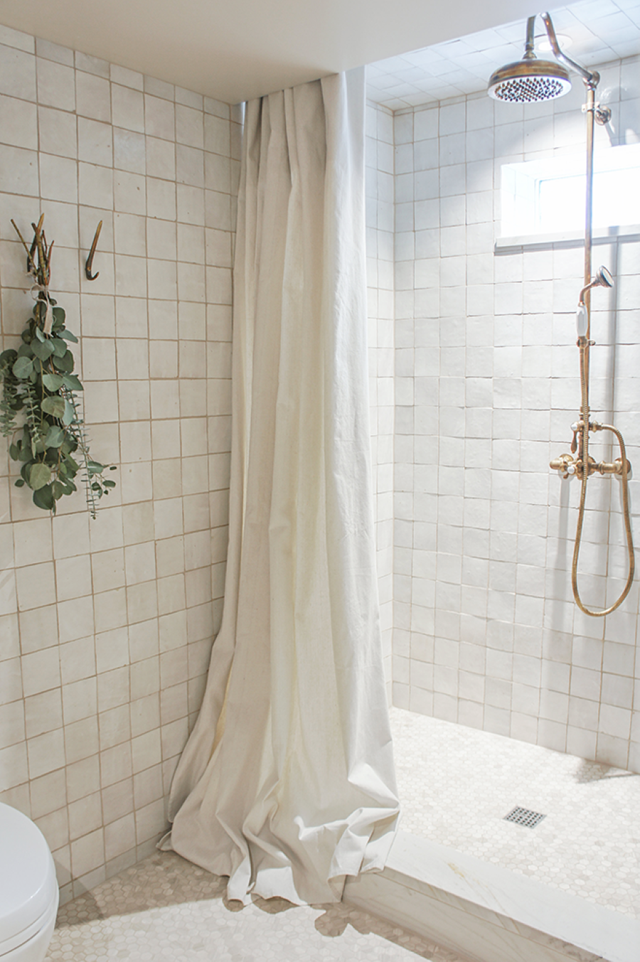 Baño moderno con azulejos cuadrados color beige. Cortina de tela blanca, larga. Grifería de cobre. Ramas de eucaliptus secas colgando.