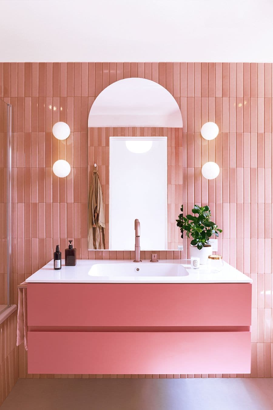 Baño con azulejos alargados color rosado. Vanitorio moderno, de color rosado y base blanca. Grifería de cobre rosa. Espejo en forma de arco. A ambos costados hay dos focos de muro redondos, uno sobre otro.