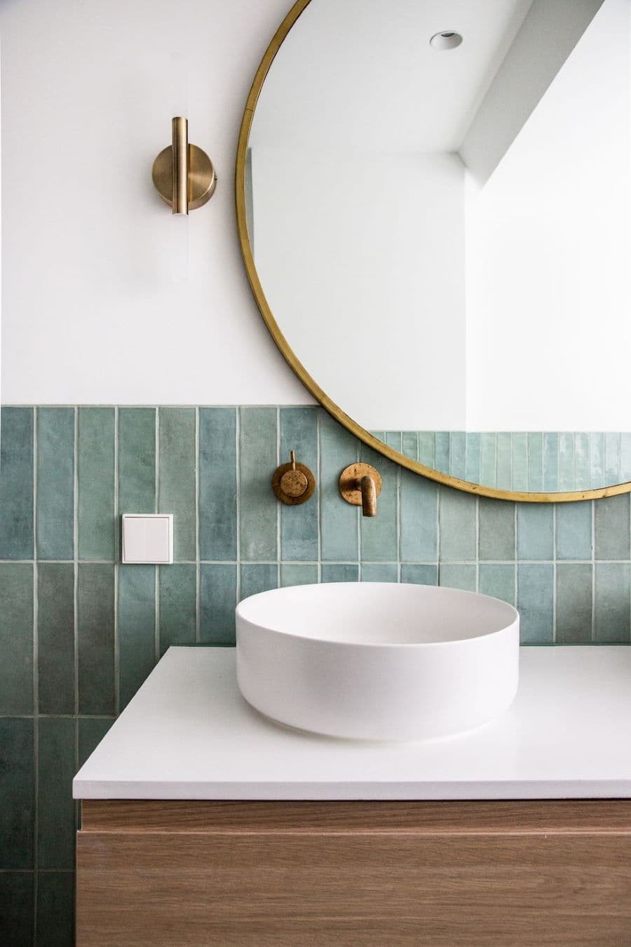 Baño con lavamanos redondo, sobrepuesto sobre un vanitorio de madera. Grifería empotrada en el muro. Azulejos azul verdoso, rectangulares. Gran espejo redondo con marco dorado.