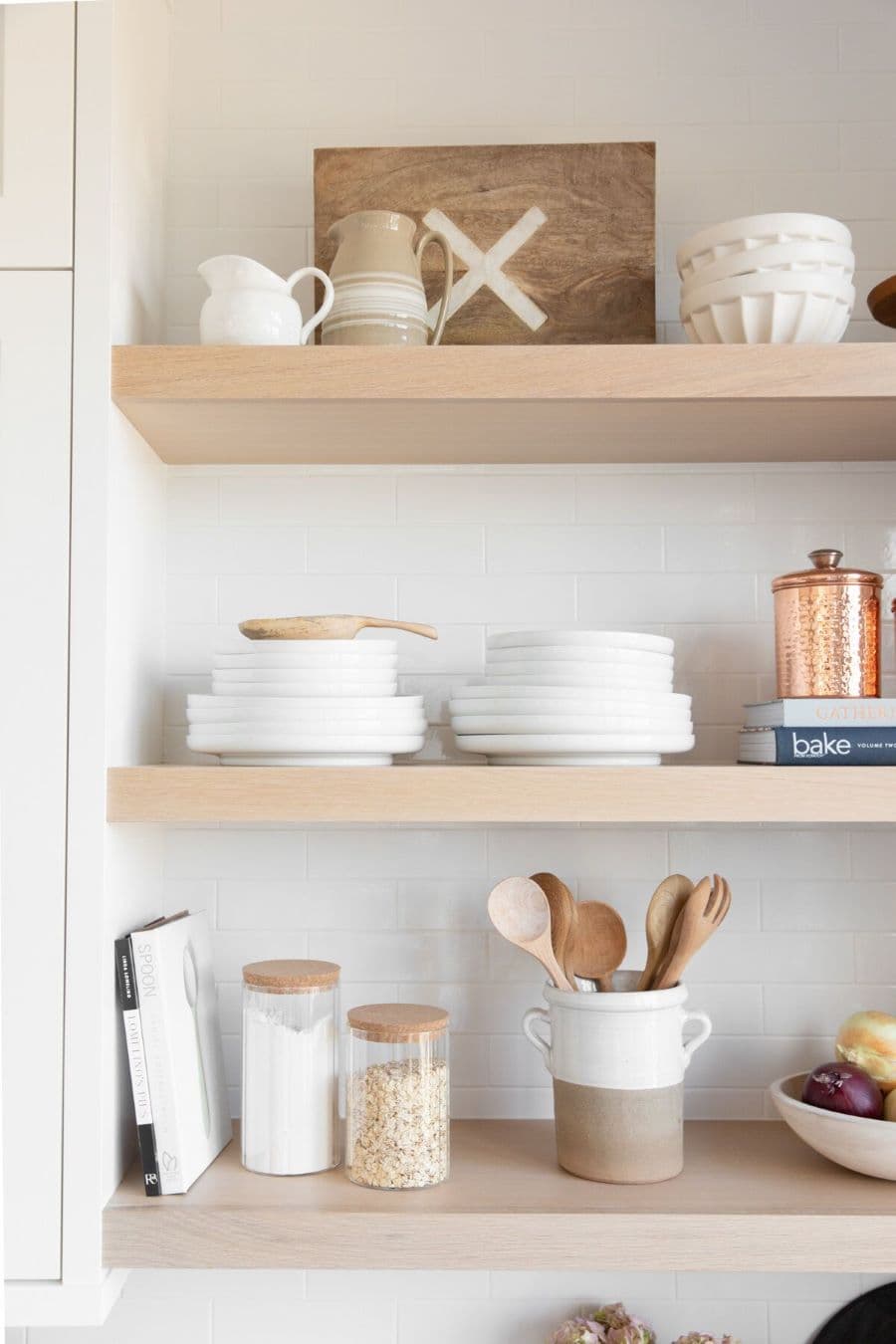 Detalle de tres repisas flotantes de una cocina. Son de madera clara y sobre ella hay vajilla de color blanco, platos, bowls, recipientes con cucharas de madera y frascos de vidrio. Muro blanco y mueble de despensa a un costado