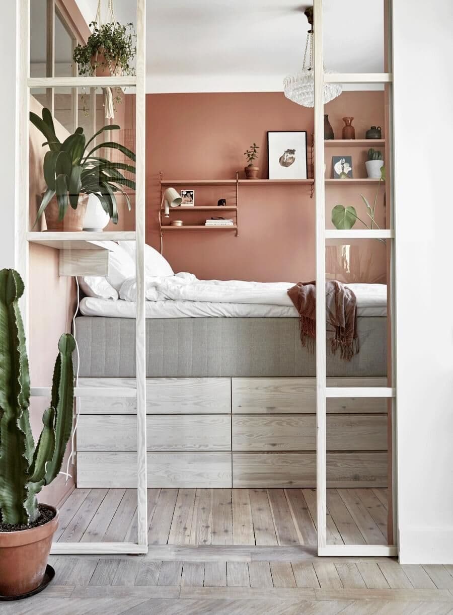 Dormitorio pequeño con muros rosa pálido y rosa oscuro. Piso de madera gris clara. La cama está en altura, sobre un mueble a medida con 6 cajones. La ropa de cama es blanca. Repisas flotantes con plantas y adornos